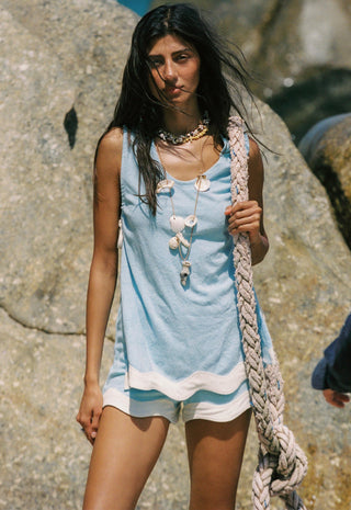 a-woman-standing-outdoors-against-large-rocks-wearing-a-light-blue-sleeveless-top-with-white-trim-and-white-shorts-holding-a-thick-braided-rope-she-is-accessorized-with-layered-shell-necklaces
