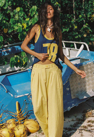 a-woman-stands-outdoors-in-front-of-a-blue-boat-and-green-foliage-wearing-a-navy-tank-top-with-yellow-trim-and-the-number-78-paired-with-loose-yellow-pants