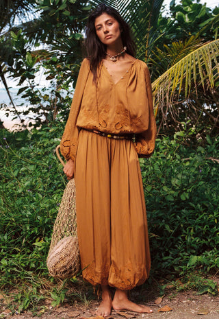 a-woman-stands-outdoors-in-a-lush-green-tropical-setting-wearing-a-matching-rust-orange-bohemian-two-piece-set-consisting-of-a-long-sleeve-embroidered-top-and-wide-leg-pants-she-is-barefoot-and-holds-a-woven-straw-bag