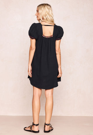 a-woman-shown-from-the-back-wearing-a-short-black-dress-with-puff-sleeves-and-a-decorative-multicolor-beaded-neckline-paired-with-black-strappy-sandals-against-a-textured-beige-studio-backdrop