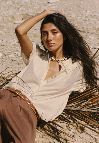 A woman reclining outdoors on a sandy ground wearing a cream linen shirt, brown pants, and a studded brown belt, accessorized with a chunky gold necklace.