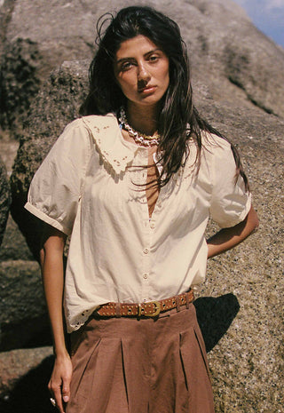 A woman standing outdoors against a rocky cliff wearing a cream short-sleeve blouse with a ruffled collar and buttons