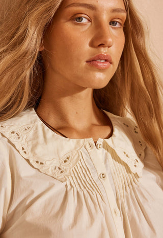 Close-up of a woman wearing a cream-colored blouse with a large eyelet lace collar and smocked chest detailing, set against a warm beige background.
