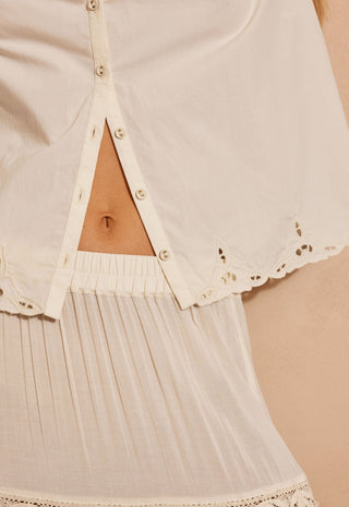 Close-up of a cream-colored eyelet lace blouse with a triangular cutout at the center front, showing the model's midriff and a pleated matching skirt.