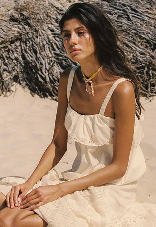 A woman sitting on a sandy beach wearing a cream-colored ruffled spaghetti-strap dress and a gold necklace.