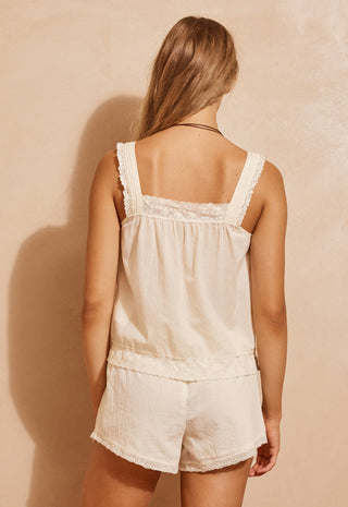 A woman shown from the back wearing a cream-colored cotton pajama set with lace trim, consisting of a sleeveless top and matching shorts, against a textured beige wall.