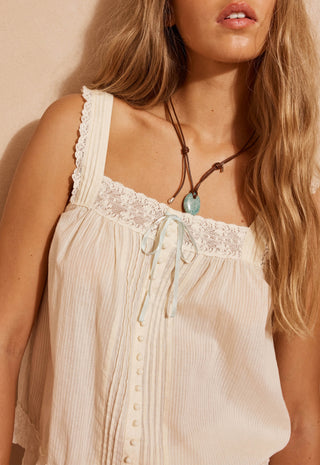 A close-up of a woman wearing a cream-colored sleeveless camisole top with lace trim, a front button placket, and a light blue ribbon tie at the neckline.