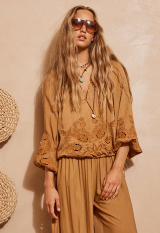 a-woman-modeling-a-bohemian-style-tan-jumpsuit-with-intricate-embroidery-and-lace-detailing-on-the-bodice-and-wide-sleeves-styled-with-layered-necklaces-and-sunglasses-against-a-textured-beige-wall