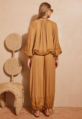 a-woman-stands-with-her-back-to-the-camera-wearing-a-camel-brown-two-piece-set-consisting-of-a-loose-long-sleeve-top-with-lace-trim-and-matching-wide-leg-pants-with-lace-detailing-at-the-hems-she-is-posed-next-to-a-woven-rattan-chair-against-a-textured-beige-wall