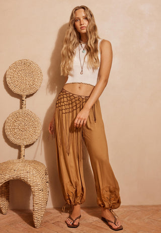 a-woman-stands-indoors-wearing-loose-tan-harem-style-pants-with-a-black-woven-waist-panel-and-drawstring-paired-with-a-white-sleeveless-crop-top-and-sandals-a-woven-chair-is-visible-at-left-against-a-warm-beige-wall