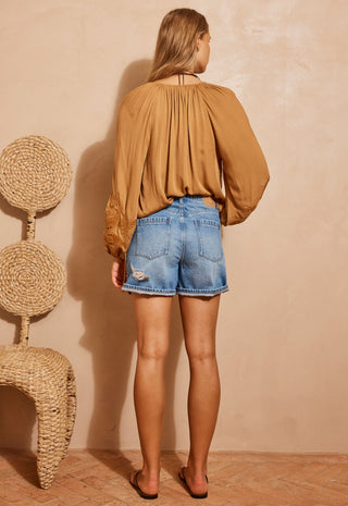 A woman stands with her back to the camera wearing a camel-colored long-sleeve blouse and light-wash denim shorts, posed in front of a textured beige wall with a woven chair on the left.