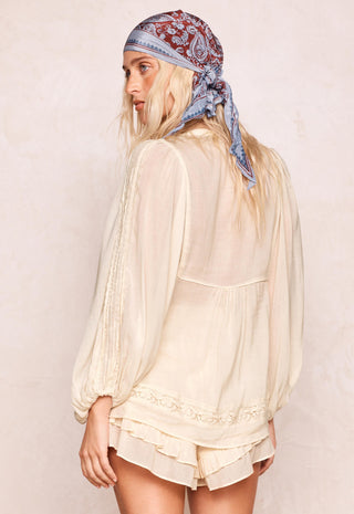 A woman shown from the back wearing a cream-colored, semi-sheer long-sleeve blouse with lace trim and a paisley bandana headscarf, set against a textured beige wall.