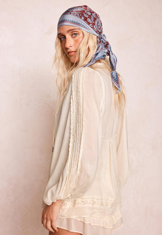 A woman wearing a cream-colored, semi-sheer long-sleeve blouse with lace trim and a blue paisley bandana headscarf, photographed against a textured beige background.