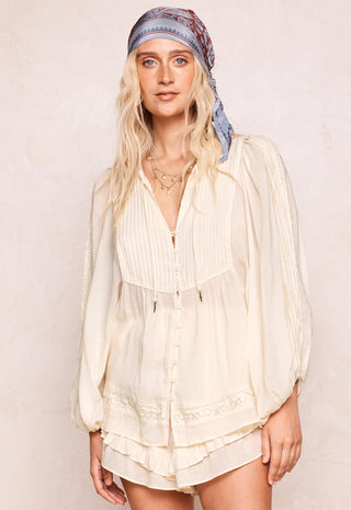 A woman wearing a cream-colored, long-sleeved bohemian-style blouse with pleated details and a ruffled hem, paired with a blue patterned headscarf, set against a neutral beige background.