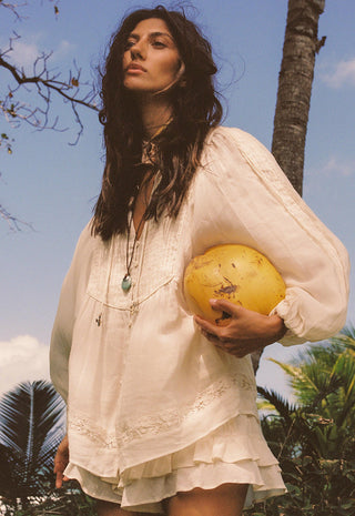 A woman outdoors wearing a loose white bohemian-style blouse and holding a yellow coconut under her arm