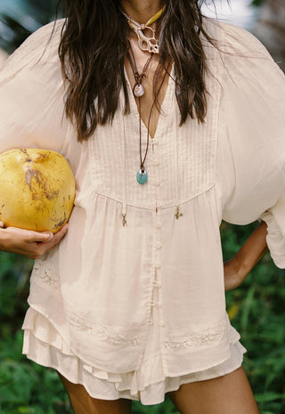 A woman wearing a cream-colored, bohemian-style short dress or romper with ruffled edges and button details, holding a yellow coconut in her hand against a green outdoor background.