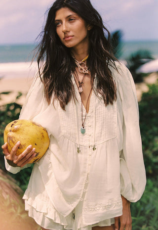 A woman on a beach wearing a loose white bohemian-style blouse and holding a yellow coconut.