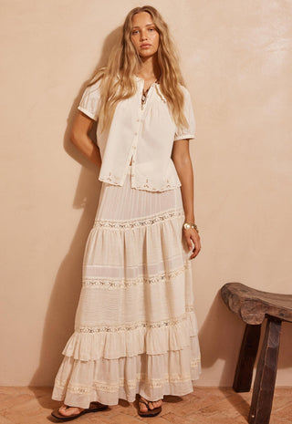 A woman modeling a bohemian-style two-piece outfit consisting of a short-sleeve cream button-up shirt and a matching long tiered maxi skirt with lace details, set against a warm beige textured wall.