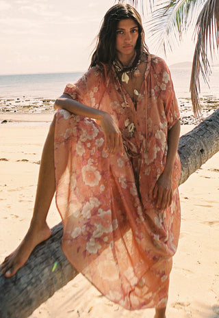 A woman on a sandy beach sits on a fallen palm tree wearing a flowing peach floral kaftan dress. The background shows the ocean