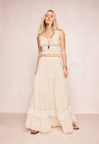 A woman modeling a cream-colored two-piece set consisting of a sleeveless crop top and a long tiered maxi skirt with lace details, set against a neutral textured beige backdrop.
