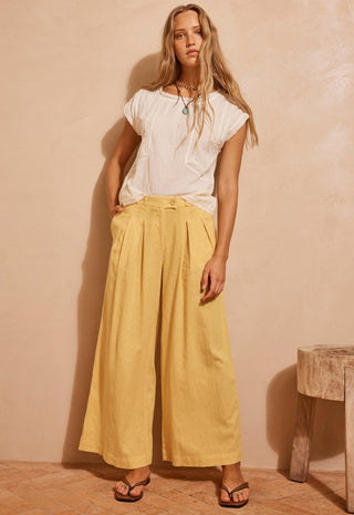 a-woman-stands-in-a-studio-wearing-a-white-short-sleeve-top-and-wide-leg-yellow-pants-with-sandals-and-a-necklace-against-a-warm-beige-wall