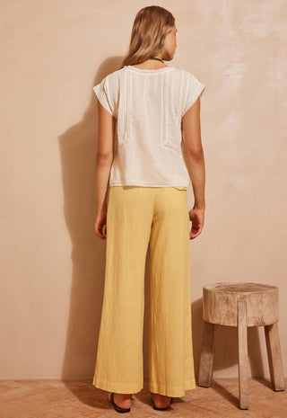 a-woman-stands-with-her-back-to-the-camera-wearing-a-cream-short-sleeve-top-and-mustard-yellow-wide-leg-pants-standing-in-front-of-a-textured-beige-wall-next-to-a-rustic-wooden-stool