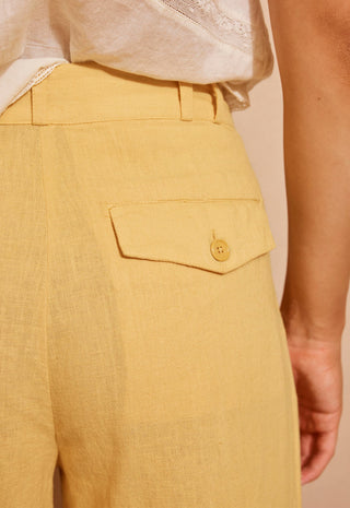 close-up-of-a-person-wearing-mustard-yellow-linen-trousers-with-a-buttoned-flap-pocket-and-a-white-top-against-a-plain-beige-background