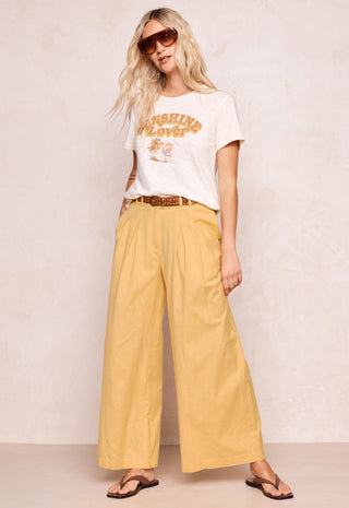 a-woman-modeling-a-white-graphic-t-shirt-and-yellow-wide-leg-pants-with-a-brown-belt-styled-with-sunglasses-and-flip-flops-against-a-textured-beige-wall