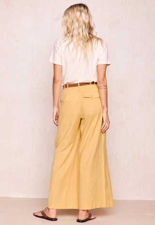 a-woman-shown-from-the-back-wearing-wide-leg-mustard-yellow-pants-a-white-t-shirt-and-brown-sandals-against-a-textured-beige-wall