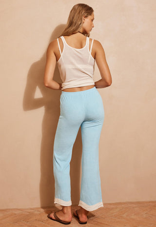 a-woman-stands-with-her-back-to-the-camera-wearing-light-blue-flared-lounge-pants-with-a-white-hem-paired-with-a-white-tank-top-and-brown-sandals-against-a-warm-beige-wall