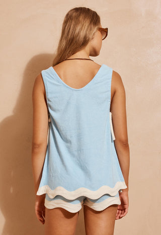 A woman shown from the back wearing a light blue sleeveless top with a scalloped white hem and matching light blue shorts, standing against a warm beige textured wall.