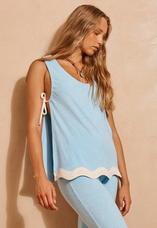 A woman wearing a light blue sleeveless knit top with white tie details on the shoulders and a white scalloped hem, paired with matching light blue pants, posing against a textured beige wall.